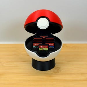 Puede incluir: Un estuche de almacenamiento esférico rojo y blanco con forma de Pokeball, que presenta una base negra y una tapa con bisagras. El interior contiene varias ranuras que albergan cartuchos de juego de Nintendo Switch con etiquetas.