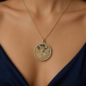 May include: A gold-toned necklace featuring a round pendant with a black recessed background. The pendant displays an embossed design of a winged figure holding a flower, surrounded by stars and a crescent moon, suspended on a thin gold chain.