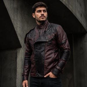 May include: A maroon and black jacket featuring a high collar, a diagonal front zipper closure, and black leather-like panels on the shoulders and sleeves. The sleeves include ribbed detailing at the elbows and cuffs, with metallic snap buttons.