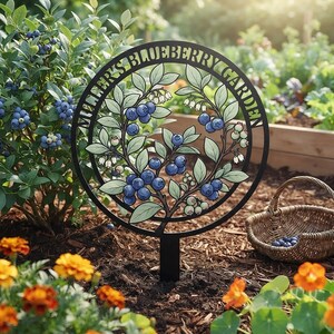 May include: A circular black metal garden sign featuring a central illustration of blueberry branches with blue berries and white flowers. The text "MILLER'S BLUEBERRY GARDEN" is engraved along the top inner rim of the frame. The sign is mounted on a stake.