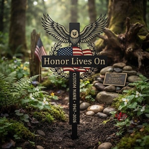May include: A black metal memorial cross stake featuring a bald eagle with spread wings perched above a waving American flag. The horizontal bar displays the text Honor Lives On, while the vertical bar includes Custom Name, 1947-2026.