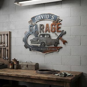 May include: A metal wall sign featuring the text DAVID'S GARAGE and BUILT FIXED RESTORED. The design includes a vintage pickup truck, a large gear, and a wrench. The sign has a weathered, multi-colored finish with blue, grey, and rust-toned accents.