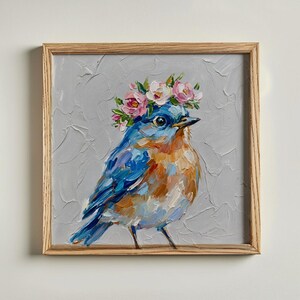 Könnte beinhalten: Ein Ölgemälde in einem hellen Holzrahmen zeigt einen blauen Vogel mit orangefarbener Brust und einem Kranz aus rosa und weißen Blumen auf dem Kopf. Der Hintergrund ist hellgrau strukturiert und der Vogel ist mit dicken Pinselstrichen gemalt.
