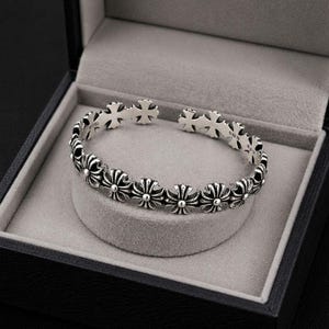 May include: A silver cuff bracelet with a repeating floral and cross pattern. The bracelet is presented in a black jewellery box with a grey velvet lining. The design of the bracelet features alternating floral and cross motifs.