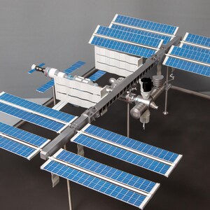 May include: A detailed scale model of the International Space Station featuring a central metallic truss structure, multiple white cylindrical modules, and several large, rectangular blue solar array wings extending from the main frame.