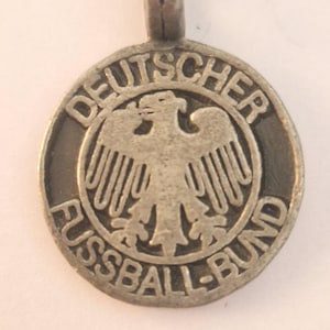 Op de afbeelding: Een ronde zilverkleurige metalen hanger met een verhoogd adelaarembleem in het midden. De tekst DEUTSCHER staat in reliëf langs de bovenrand en de tekst FUSSBALL-BUND staat in reliëf langs de onderrand van de cirkelvormige lijst.