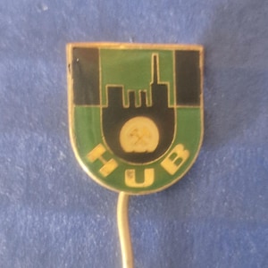 May include: A vintage pin with a shield shape. The pin features a green and black design with a stylized cityscape and the letters "HUB" in gold. The pin has a gold-colored metal border and a pin backing.
