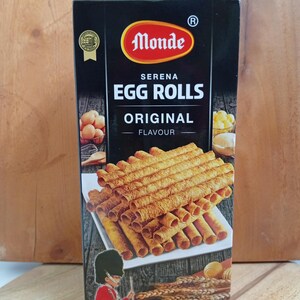 May include: A rectangular black box of Monde Serena Original Flavour Egg Rolls. The packaging features a stack of golden-brown cylindrical wafer rolls, images of eggs, wheat stalks, and butter, and a graphic of a soldier in a red uniform and bearskin hat.