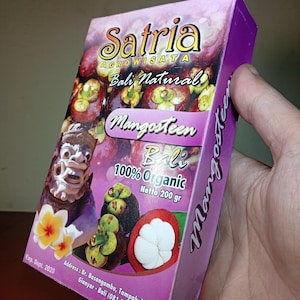 Pode incluir: Uma caixa retangular roxa de suplemento de mangostão Satria Agrowisata Bali Natural. A embalagem apresenta imagens de mangostão, uma máscara balinesa e flores de frangipani amarelas. Texto: 100% Organic, Netto 200 gr, Exp. Sept. 2028.