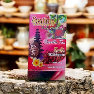 Puede incluir: Caja rosa de té Rosella Satria de Bali, 100% orgánico. El empaque presenta texto e ilustraciones, incluyendo un templo balinés y una flor. El peso neto es de 100 gramos.