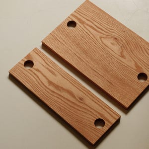 Può includere: Due taglieri rettangolari in legno con venature naturali. Ogni tagliere ha un foro circolare in un angolo. Il legno ha una calda tonalità marrone-rossastra. I taglieri sono di dimensioni diverse.