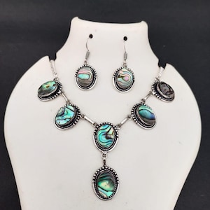 May include: A jewelry set consisting of a necklace and matching drop earrings. Each piece features oval-shaped abalone shell inlays with iridescent green, blue, and pink tones, set in silver-toned metal frames with a beaded, rope-like texture.
