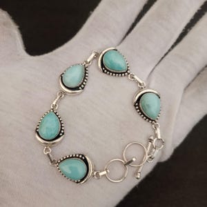 Peut inclure: Un bracelet en argent composé de cinq pierres bleu clair en forme de goutte, serties dans des montures métalliques décoratives. Les pierres sont reliées par des maillons en argent et le bijou se ferme par un fermoir à bascule avec anneau.