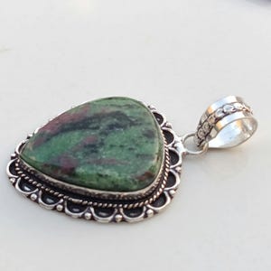 Puede incluir: Un colgante en forma de lágrima con una piedra pulida de color verde y rojo oscuro, engastada en un marco de plata decorativo con borde festoneado y detalle de cuerda trenzada, con una anilla cilíndrica de plata en la parte superior.