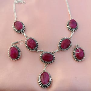 May include: A silver necklace and earring set featuring oval-shaped, deep red gemstones. The gemstones are set in a silver frame with a beaded border. The necklace has a chain and the earrings have a hook closure. The jewelry set is displayed on a pink surface.