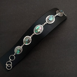 Könnte beinhalten: Ein silberfarbenes Gliederarmband mit fünf ovalen Abalone-Muscheleinlagen in schillernden Grün-, Blau- und Violetttönen. Das Armband besteht aus einer Reihe kreisförmiger Metallglieder und einem kleinen Verschlussmechanismus an einem Ende der Kette.