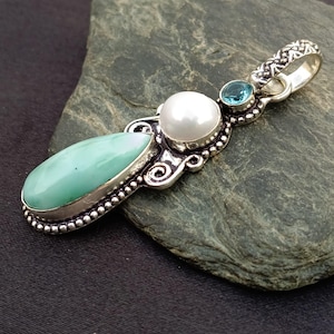 May include: A silver pendant featuring a teardrop-shaped light green stone, a round white pearl, and a small round blue gemstone. The metal setting has a beaded edge and a textured bail at the top, showcasing intricate metalwork and polished stones.
