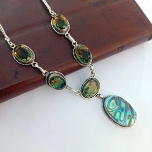 Puede incluir: Un collar de plata con piedras preciosas ovaladas verdes y amarillas y un colgante de abulón. El collar tiene una cadena delicada y está sobre un fondo marrón. El colgante de abulón tiene un patrón iridiscente azul y verde.
