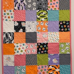 May include: A patchwork quilt with a pink border. The quilt is composed of numerous squares, each featuring a different Halloween-themed pattern, including bats, skulls, spiderwebs, and ghosts. The color palette includes orange, black, pink, and purple.