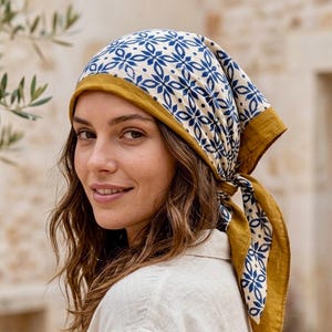 May include: A square headscarf featuring a white base with a repeating blue floral pattern and a solid mustard yellow border. The fabric is tied at the back of the head, with the mustard yellow edges framing the face and the patterned section covering the top.