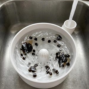 Può includere: Una bacinella circolare bianca con una cupola centrale e un beccuccio laterale, riempita d'acqua e oggetti rotondi scuri. Un getto d'acqua scorre da un rubinetto nel beccuccio laterale, creando un movimento vorticoso all'interno della bacinella.