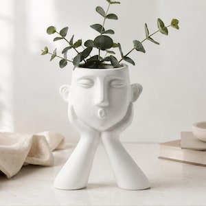 Puede incluir: Una maceta de cerámica blanca mate con forma de cabeza humana que descansa sobre dos manos. El rostro presenta ojos cerrados y labios fruncidos. Una planta verde con pequeñas hojas redondas está colocada en la abertura superior del recipiente.