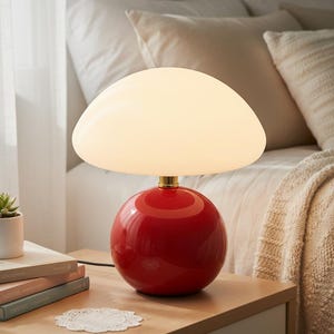 May include: A red, spherical table lamp with a white, mushroom-shaped shade. The lamp sits on a wooden nightstand next to a small potted plant and a stack of books. The lamp has a gold-colored base.