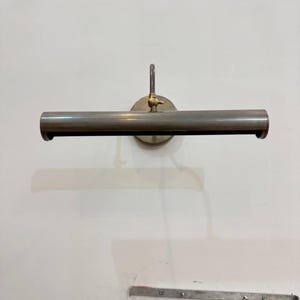 May include: A wall-mounted picture light with a long, horizontal, cylindrical metal shade in a brushed nickel finish. The fixture features a central brass-toned adjustment knob and a circular mounting plate, designed to illuminate artwork.