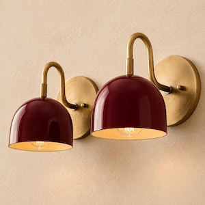 May include: Two wall sconces featuring glossy, deep red, dome-shaped shades attached to curved, brushed brass arms. Each lamp is mounted on a circular brass backplate, with a single light bulb visible inside the open bottom of the red metal shade.