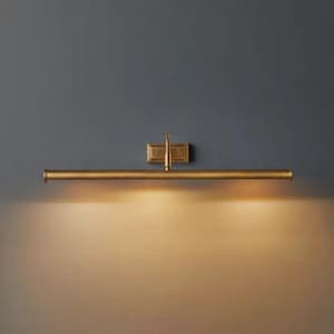 May include: A horizontal wall-mounted picture light fixture with a metallic brass finish. The design features a long, slender cylindrical tube attached to a rectangular wall plate by a central vertical arm, emitting a warm downward glow.