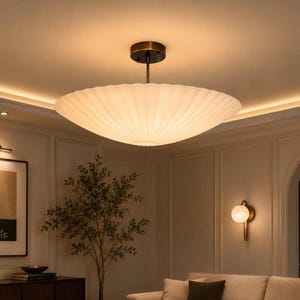 May include: A large, circular, white, pleated, saucer-shaped ceiling light fixture with a dark bronze-colored metal stem and canopy. The light emits a soft, warm glow, illuminating the room from a central position on the ceiling.