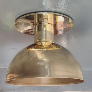 May include: A polished brass flush mount ceiling light fixture featuring a dome-shaped shade attached to a circular base plate. The metal surface has a reflective finish with a visible knurled screw detail on the central cylindrical neck.