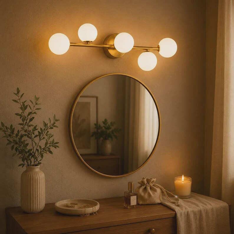 May include: A wall-mounted vanity light fixture with a brass finish and four spherical white glass globes. The light is positioned above a circular mirror with a thin brass frame, reflecting a room interior with a plant and furniture.