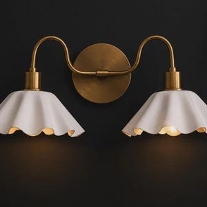 Può includere: Una lampada da parete doppia con una piastra circolare in ottone e due bracci curvi in ottone. Ogni braccio sostiene un paralume bianco a forma di campana con bordi ondulati, da cui emana una luce calda e soffusa.