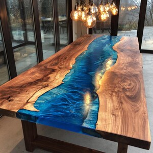 May include: A large, rectangular wooden table with a striking blue epoxy resin river running through the center. The table's edges are natural wood, showcasing the grain. The table is supported by dark brown wooden legs.