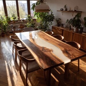 Op de afbeelding: Een grote, rechthoekige houten eettafel met een lichtgekleurde epoxyharsrivier in het midden. De tafel is omringd door houten stoelen met grijze gestoffeerde zittingen. De kamer heeft een warme sfeer met natuurlijk licht en kamerplanten.