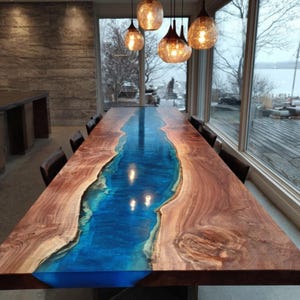 May include: A large, rectangular dining table with a unique design. The table features a river of blue epoxy resin flowing through the center, framed by natural wood edges. The table is surrounded by chairs and lit by hanging globe lights.