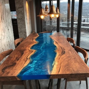 May include: A large, rectangular wooden dining table with a blue epoxy resin river running down the center. The table is surrounded by wooden chairs and illuminated by hanging pendant lights. The table is approximately 100 inches long.