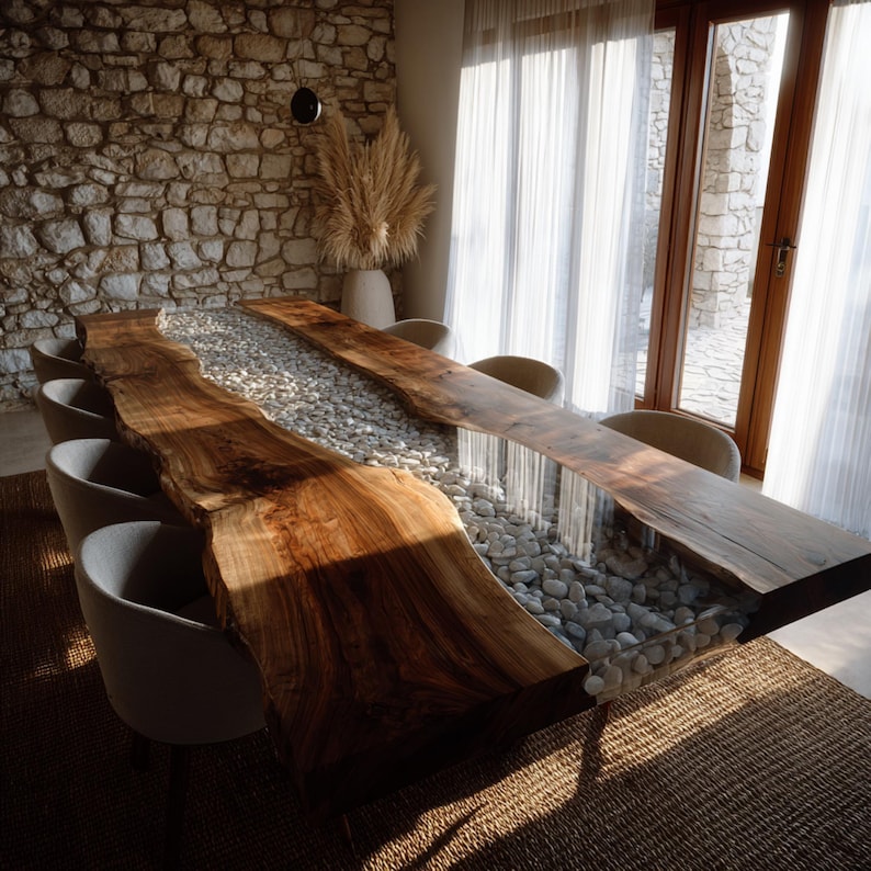 May include: A bespoke wooden dining table with a central river of white pebbles. The table has a live edge design and is surrounded by neutral chairs. The room features a stone wall and large windows, creating a natural aesthetic.