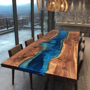 May include: A large, rectangular wooden dining table with a blue resin river design. The table is surrounded by eight chairs with brown upholstery. The table is made of natural wood with a glossy finish. The room has large windows and a modern chandelier.