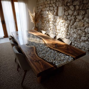 May include: A bespoke dining table with a live edge design. The dark wood table has a clear resin river filled with white pebbles. The table is surrounded by upholstered chairs. The room has a stone wall and a natural, rustic aesthetic.