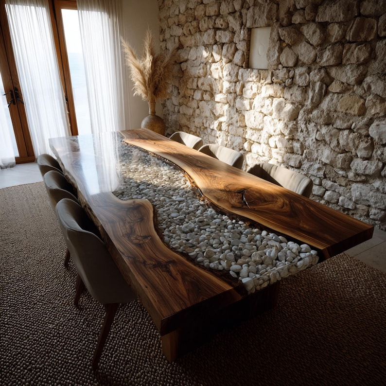 May include: A bespoke dining table with a live edge design. The dark wood table has a clear resin river filled with white pebbles. The table is surrounded by upholstered chairs. The room has a stone wall and a natural, rustic aesthetic.