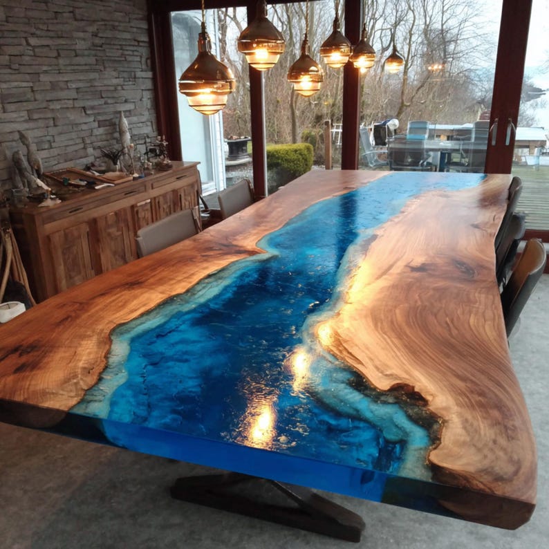 May include: A large, custom-made dining table with a unique design. The table features a wooden surface with a river of blue epoxy resin running through the center. The wood has a natural, warm brown tone, and the resin is a vibrant blue. The table is illuminated by hanging lights.