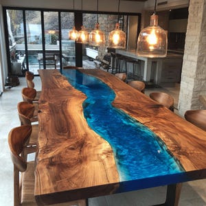 May include: A large, handcrafted dining table with a river of blue epoxy resin flowing through the center. The table is made of natural wood with a live edge, and measures approximately 120 inches long. Wooden chairs surround the table.