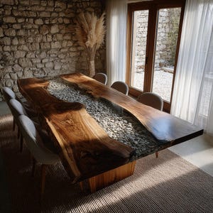 May include: A bespoke dining table with a live edge design. The table has a central river of smooth stones set in clear resin. The wooden tabletop is a dark brown colour, and the base is made of wood. The table is surrounded by grey chairs. The table is approximately 300 cm long.