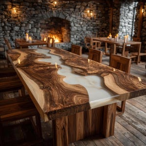 May include: A large, handcrafted wooden dining table with a river-like epoxy resin center. The table features a natural wood grain pattern with a light-colored resin. The table is supported by a sturdy wooden base and surrounded by wooden chairs.