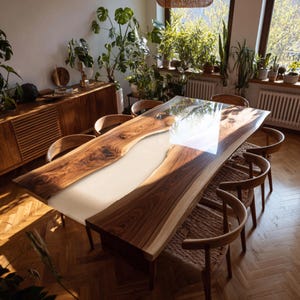 Op de afbeelding: Een grote, op maat gemaakte eettafel met een live edge design. De tafel heeft een lichtgekleurd epoxyhars middenstuk en is omgeven door natuurlijk hout. De tafel wordt gecombineerd met houten stoelen en staat op een geweven vloerkleed. De kamer is gevuld met planten.