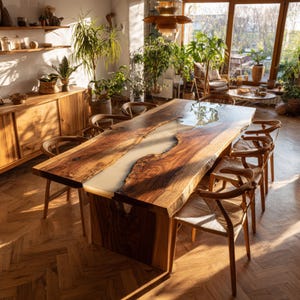 Op de afbeelding: Een grote, handgemaakte houten eettafel met een lichtgekleurde harsrivier in het midden. De tafel is omringd door houten stoelen met geweven zittingen, geplaatst in een zonnige kamer met planten.