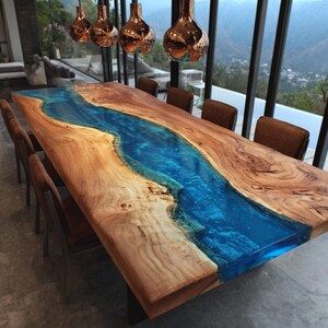 May include: A large wooden dining table with a river of blue resin running down the center. The table is surrounded by brown chairs. Copper-colored pendant lights hang above the table, and a scenic view is visible through the windows.