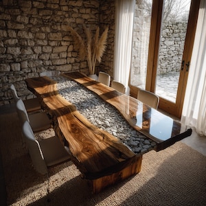 May include: A bespoke wooden dining table with a live edge design. The table has a central river of smooth, grey stones set in clear resin. The table is surrounded by cream-colored chairs and sits on a woven rug. The table measures approximately 300 cm in length.